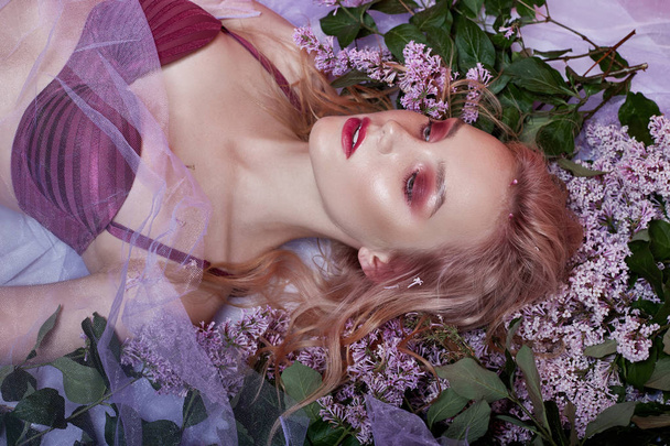 Portrait of sexy blonde in lingerie with bright makeup lying in lilac flower. Slender blonde, perfect clean skin and long hair. Natural cosmetics for skin care. Branches of lilac Bush near woman face - Foto, Bild