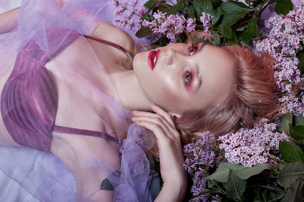Portrait of sexy blonde in lingerie with bright makeup lying in lilac flower. Slender blonde, perfect clean skin and long hair. Natural cosmetics for skin care. Branches of lilac Bush near woman face - 写真・画像