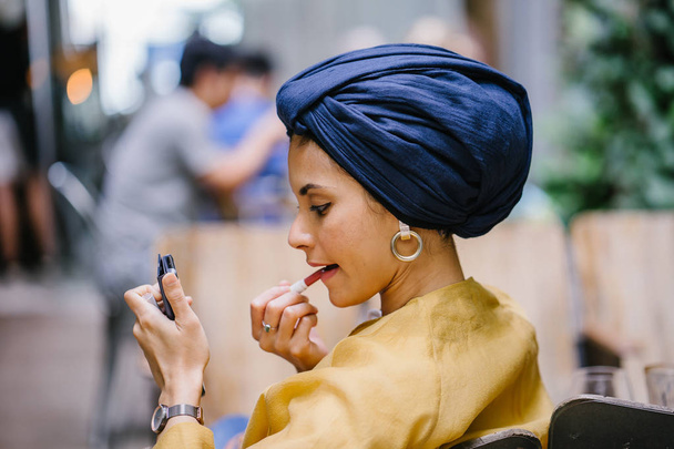 A stylish and young Muslim Malay woman wearing a turban (hijab, head scarf) applying lipstick and makeup in a cafe during the daytime. She is attractive, elegant and fashionably dressed. - Foto, Bild