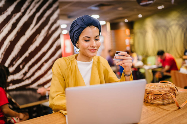 Portrait of a young and attractive Malay Muslim woman wearing a turban (hijab, head scarf). She is holding her credit card and keying in details to buy something online. - Photo, Image
