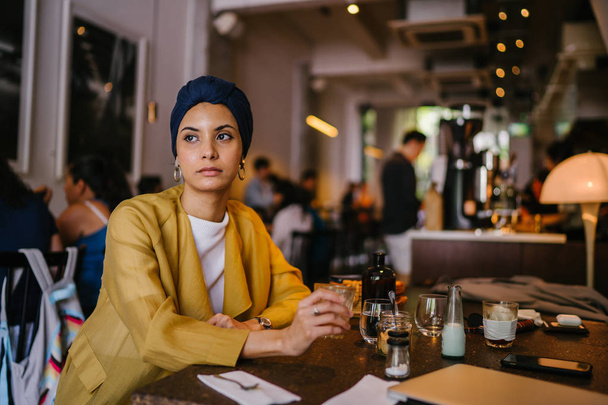 young and elegant Muslim Malay woman with  her coffee in a cafe. She is wearing a stylish outfit and turban (hijab, head scarf) and earrings. - Foto, Imagen
