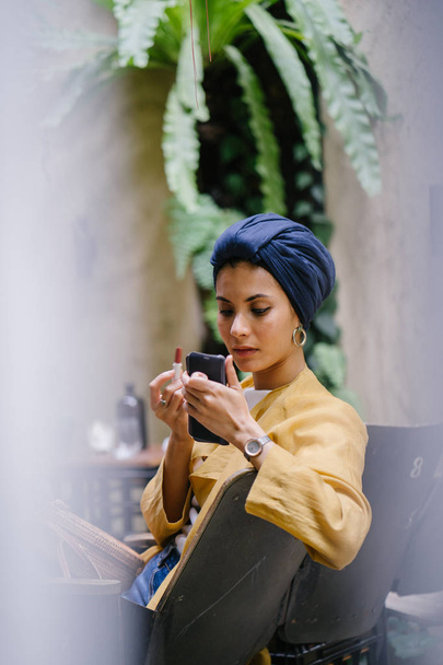 A stylish and young Muslim Malay woman wearing a turban (hijab, head scarf) applying lipstick and makeup in a cafe during the daytime. She is attractive, elegant and fashionably dressed. - Foto, Imagem