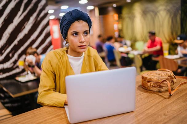 Portrait of a young Muslim Malay woman working or studying on her laptop computer indoors. She is dressed in a modest by stylish pastel outfit and a turban (hijab, head scarf). - Foto, Bild