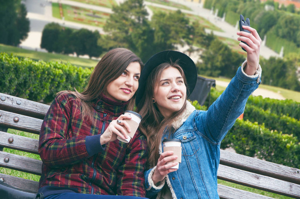 Beautiful European girls drink coffee and make selfie on the bench against the backdrop of the city - Photo, image