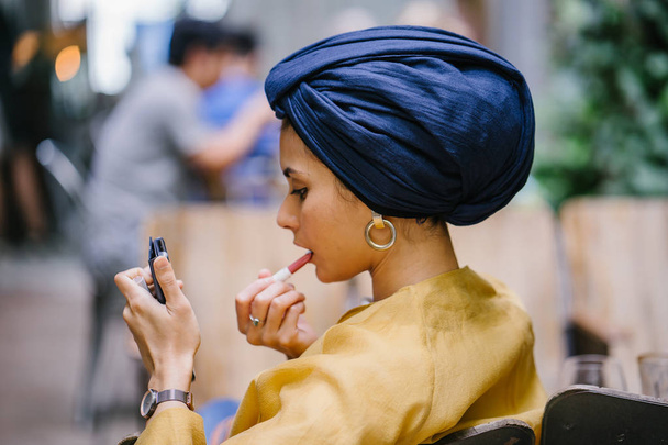 A stylish and young Muslim Malay woman wearing a turban (hijab, head scarf) applying lipstick and makeup in a cafe during the daytime. She is attractive, elegant and fashionably dressed. - Фото, зображення