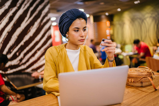 Portrait of a young and attractive Malay Muslim woman wearing a turban (hijab, head scarf). She is holding her credit card and keying in details to buy something online. - Foto, Bild