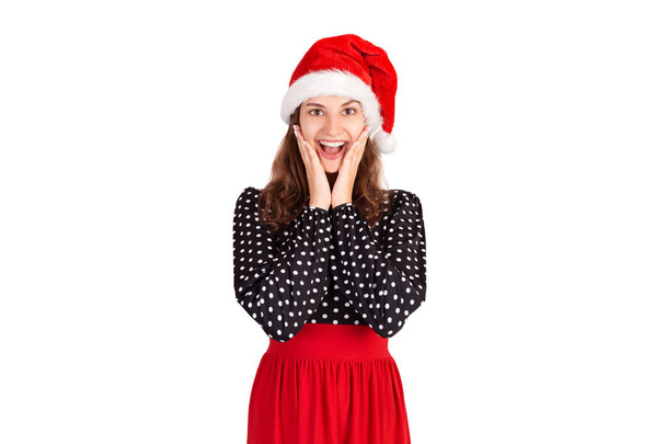 Frau im Kleid, die hochgehoben und berührt wird, die Hände vor offenen Augen hält und breit lächelt. emotionale Mädchen in Weihnachtsmann Weihnachtsmütze isoliert auf weißem Hintergrund. Urlaubskonzept. - Foto, Bild