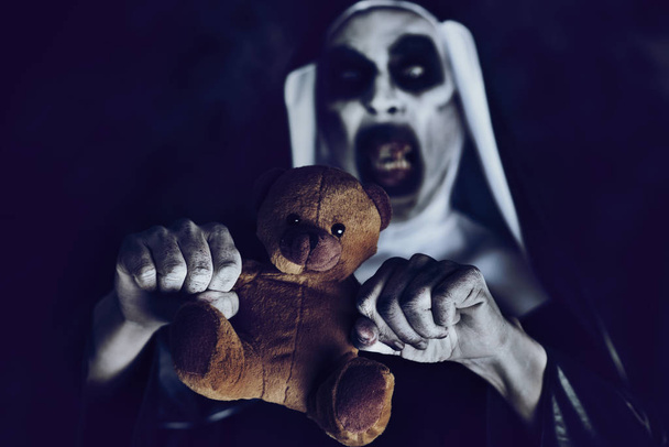 closeup of a frightening evil nun, with bloody teeth and scary eyes, wearing a typical black and white habit, trying to dismember a teddy bear against a black background - Photo, Image