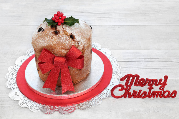 italienische Panettone-Torte mit roter Glitzerschleife und fröhlichem Weihnachtsschild mit Winterholzstechpalme auf rustikalem Holzhintergrund. traditionelles süßes Brot. - Foto, Bild