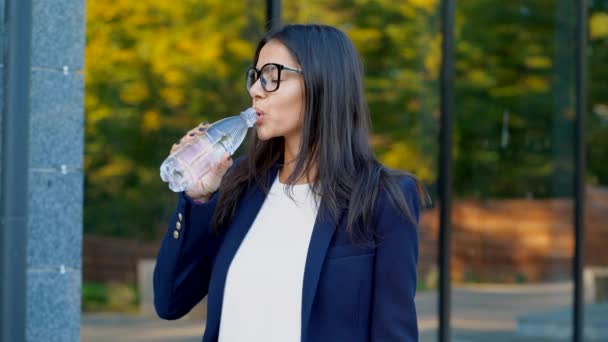 Young businesswoman near office building drinks mineral water from bottle. Beautiful girl in eyeglasses drinking clean water. Healthy lifestyle concept - Video, Çekim