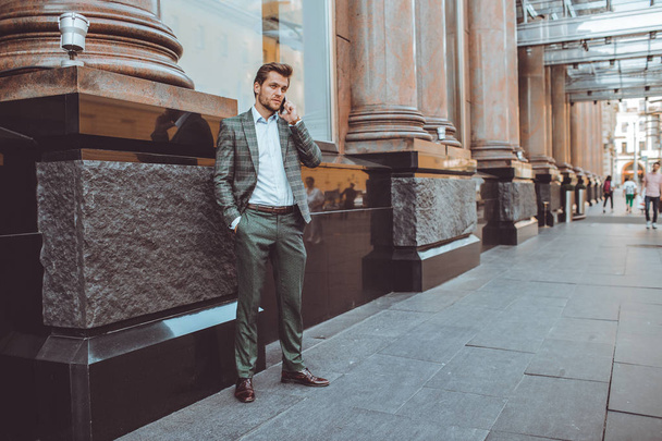 serious handsome Caucasian businessman talking on mobile phone while standing in city street at building - Фото, изображение
