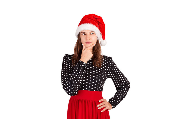 Dreamy attractive, thinking and confident young woman in dress, holding palm on cheek and gazing at upper left corner. emotional girl in santa claus christmas hat isolated on white background. - Photo, Image