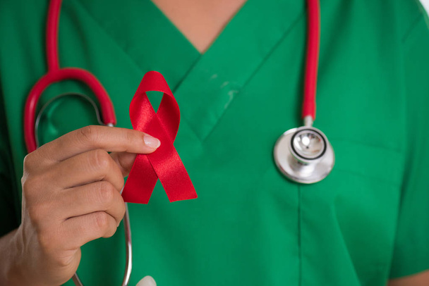 Female doctor with a stethoscope on shoulder holding red ribbon HIV, world AIDS day awareness ribbon. Healthcare and medicine concept. - Foto, Imagen