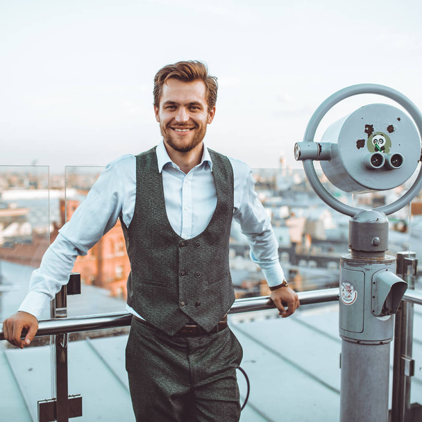 smiling stylish Caucasian man in suit Vest standing at city view telescope on roof  - Fotoğraf, Görsel