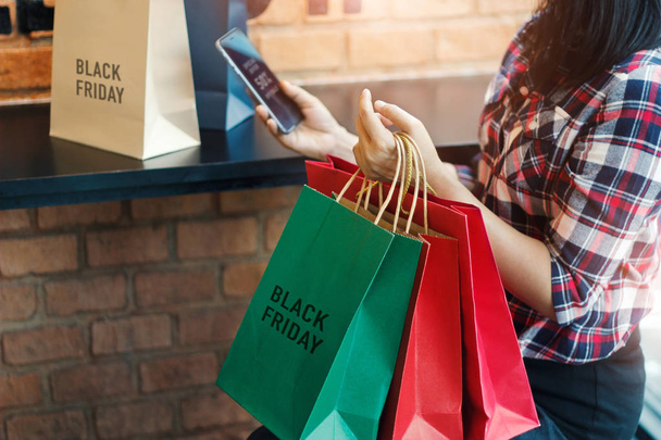 Viernes Negro, Mujer usando un teléfono inteligente y sosteniendo la bolsa de compras mientras está sentado en el fondo del centro comercial
 - Foto, imagen