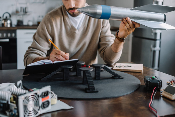 cropped image of engineer holding rocket model and making notes at home - Foto, Bild