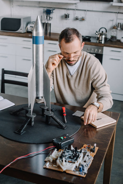 ingeniero guapo modelado cohete y mirando por encima de las gafas en la mesa en casa
 - Foto, imagen