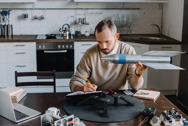 handsome engineer holding rocket model and making notes at home - Photo, Image