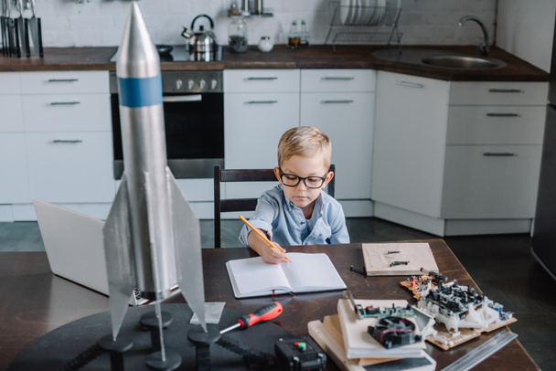 adorable boy modeling rocket and writing something to notebook in kitchen on weekend - Photo, Image