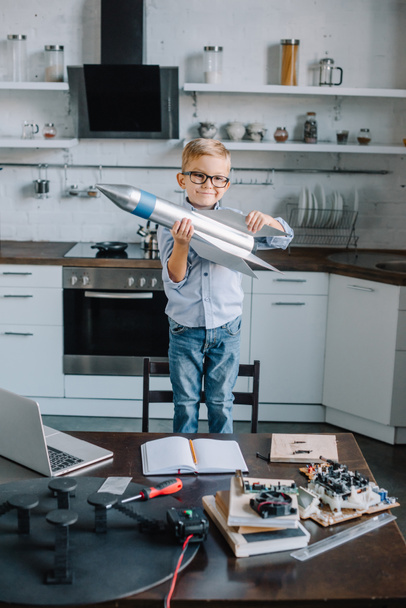 adorable boy holding rocket model in kitchen and looking at camera - Foto, imagen