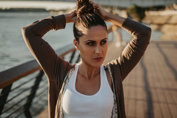 Beautiful serious woman with green eyes fixing hair bun by the river. Sunny day - Photo, Image