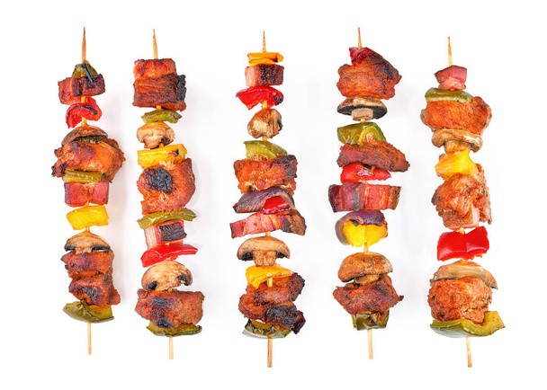 Roasted pork skewers with bacon, diced red, yellow, green bell pepper, champignons and onion stuffed on wooden sticks. Colorful mix of tasty pork barbecue on white background - Photo, Image
