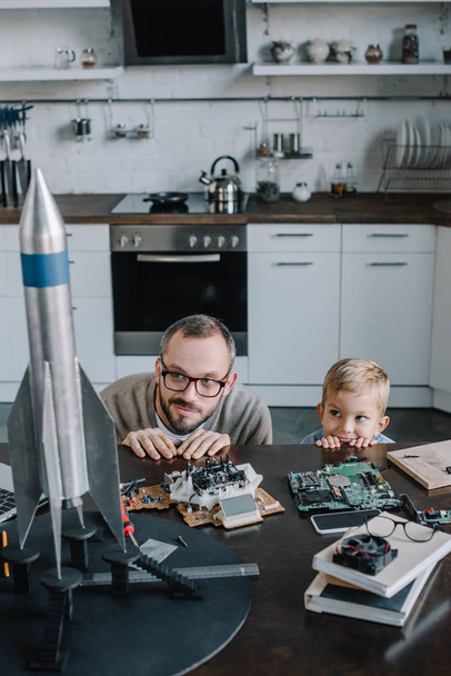 father and son looking out from table in kitchen and looking at rocket model - Photo, Image