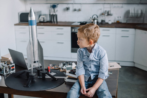 irritated grimacing boy sitting on table near rocket model in kitchen on weekend - Photo, Image