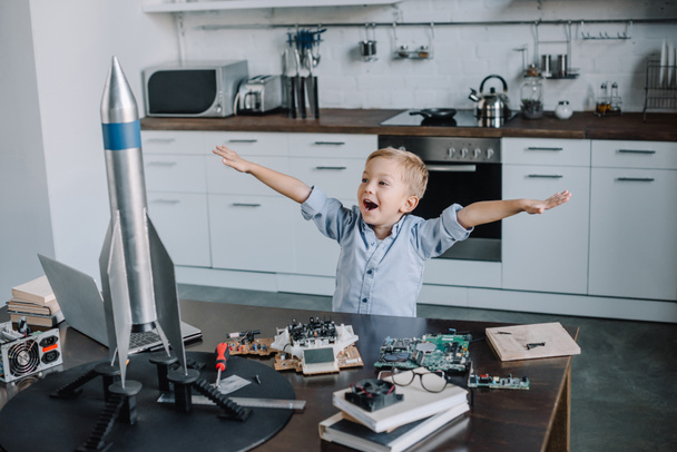 happy adorable boy standing with outstretched hands near rocket model in kitchen on weekend - Photo, Image