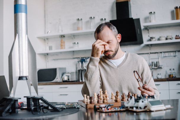 tired handsome man playing chess and touching nose bridge at home - Photo, Image
