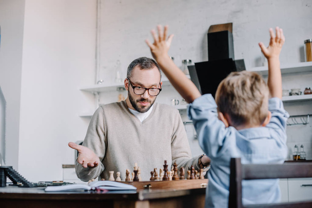 triumphing little son raising hands while playing chess with father at home - Фото, зображення