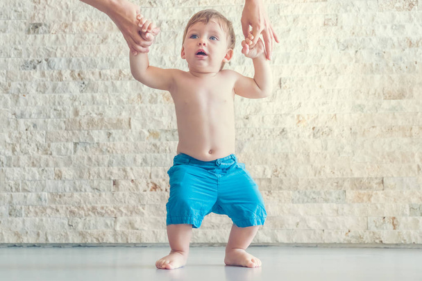 Cute blond blue-eyed baby boy in blue shorts making the first steps with the help of the mother - 写真・画像