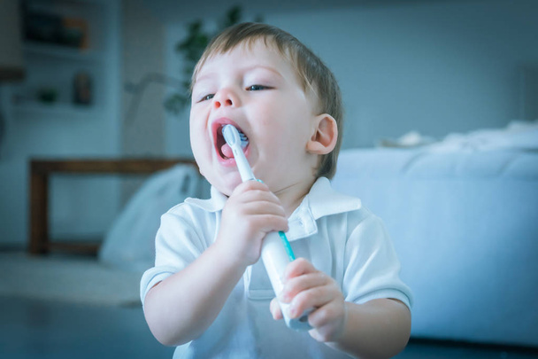 Cute small blue-eyed blond boy in a white polo shirt brushing his teeth with a toothbrush - Photo, image