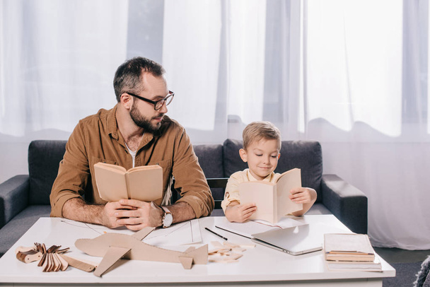 father and little son holding books while modeling plane at home - Фото, изображение