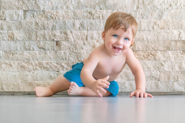 Cute laughing blue-eyed baby boy in blue shorts crawling on the floor in the room - Fotoğraf, Görsel