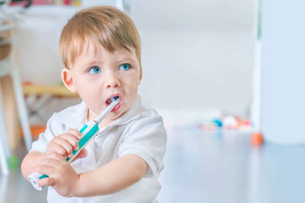Cute small blue-eyed blond boy in a white polo shirt brushing his teeth with a toothbrush - Fotografie, Obrázek