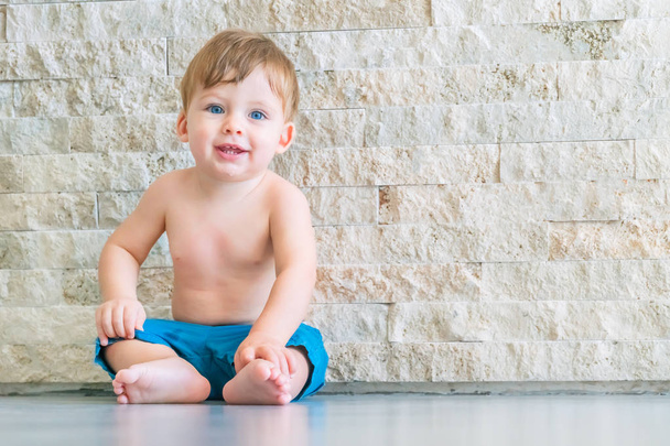 Blue-eyed blond baby boy in blue shorts sitting on the floor near a stone wall - 写真・画像