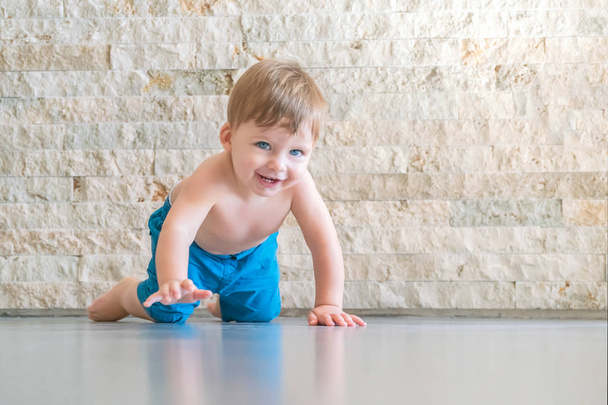 Cute laughing blue-eyed baby boy in blue shorts crawling on the floor in the room - Zdjęcie, obraz