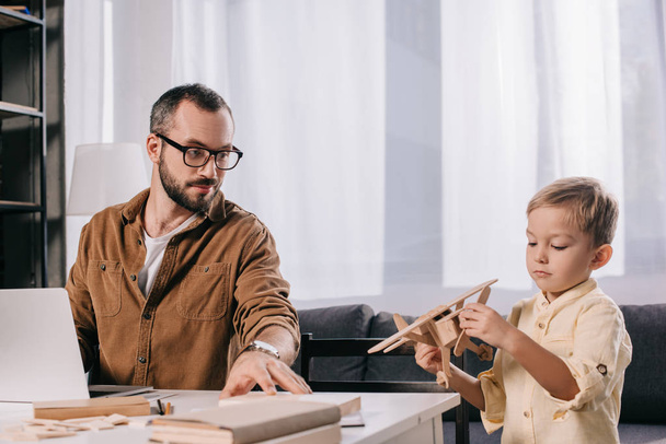 ο πατέρας που χρησιμοποιούν φορητό υπολογιστή ενώ αξιολάτρευτο μικρό γιο παίζοντας με ξύλινα αεροπλάνο μοντέλο στο σπίτι  - Φωτογραφία, εικόνα