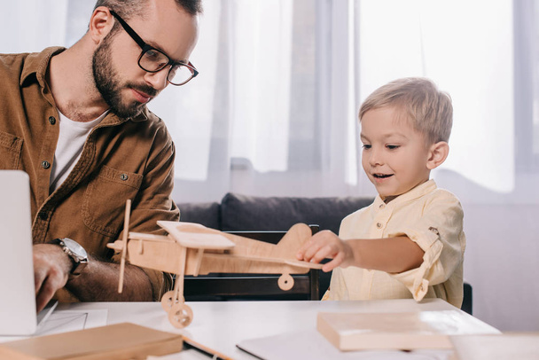笑顔の息子は木製の飛行機模型で遊んでいる間ノート パソコンを使用しての父  - 写真・画像