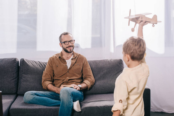 glücklicher Vater sitzt auf Couch und schaut Sohn beim Spielen mit Holzspielzeug an  - Foto, Bild