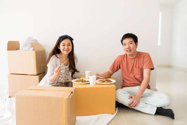 Feliz pareja china asiática sentada en el suelo y comiendo fideos de huevo de pato asado en cajas móviles de cartón en una casa nueva. Comprar una casa nueva para comenzar la vida familiar
. - Foto, imagen