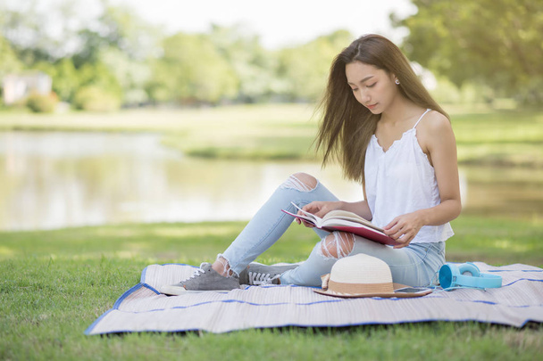 Portrait of bohemian Asian beautiful woman read text book and sit on mat at field near pond. Outdoor education lifestyle concept. Brunette woman in summer park. - Photo, Image