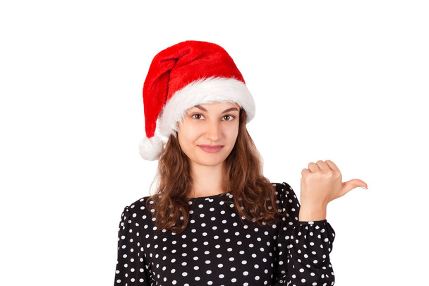 portrait of woman in dress pointing left with thumb and confused. emotional girl in santa claus christmas hat isolated on white background. holiday concept. - Фото, зображення