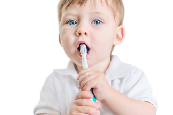 Cute small blue-eyed blond boy in a white polo shirt brushing his teeth with a toothbrush on a white background. Isolated - Foto, imagen