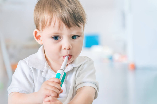 Cute small blue-eyed blond boy in a white polo shirt brushing his teeth with a toothbrush - Valokuva, kuva