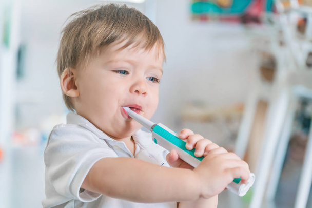 Cute small blue-eyed blond boy in a white polo shirt brushing his teeth with a toothbrush - Foto, afbeelding
