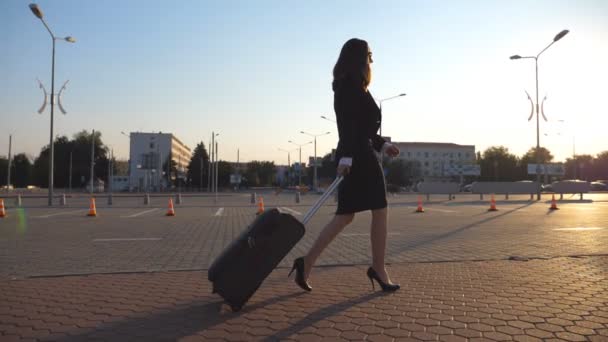 Деловая женщина с чемоданом идет в аэропорт в командировку. Дама в туфлях на высоких каблуках шагает со своим багажом вдоль улицы на закате. Концепция путешествия. Вид сбоку Медленное движение
 - Кадры, видео