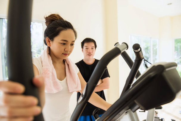 Asian woman in sports clothing in gym doing cardio workout, exercising on recumbent bicycle with her personal trainer. Sport fitness and healthy lifestyle concept. - Photo, Image