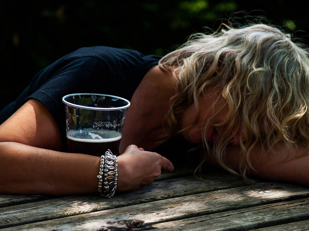 eine betrunkene Frau mit einem Glas Bier in der Hand, bewusstlos, an einen Tisch gelehnt - Foto, Bild